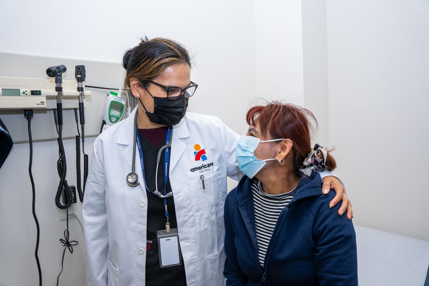 nurse on left in white lab coat stand with arm around patient in an examination room